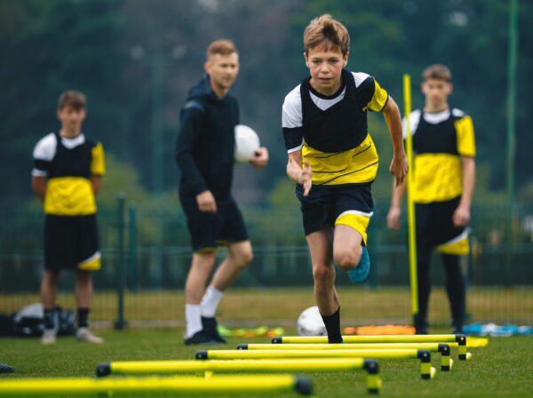 A young boy in sportswear runs through a ladder drill on a grassy field, while three others watch in the background holding soccer balls.