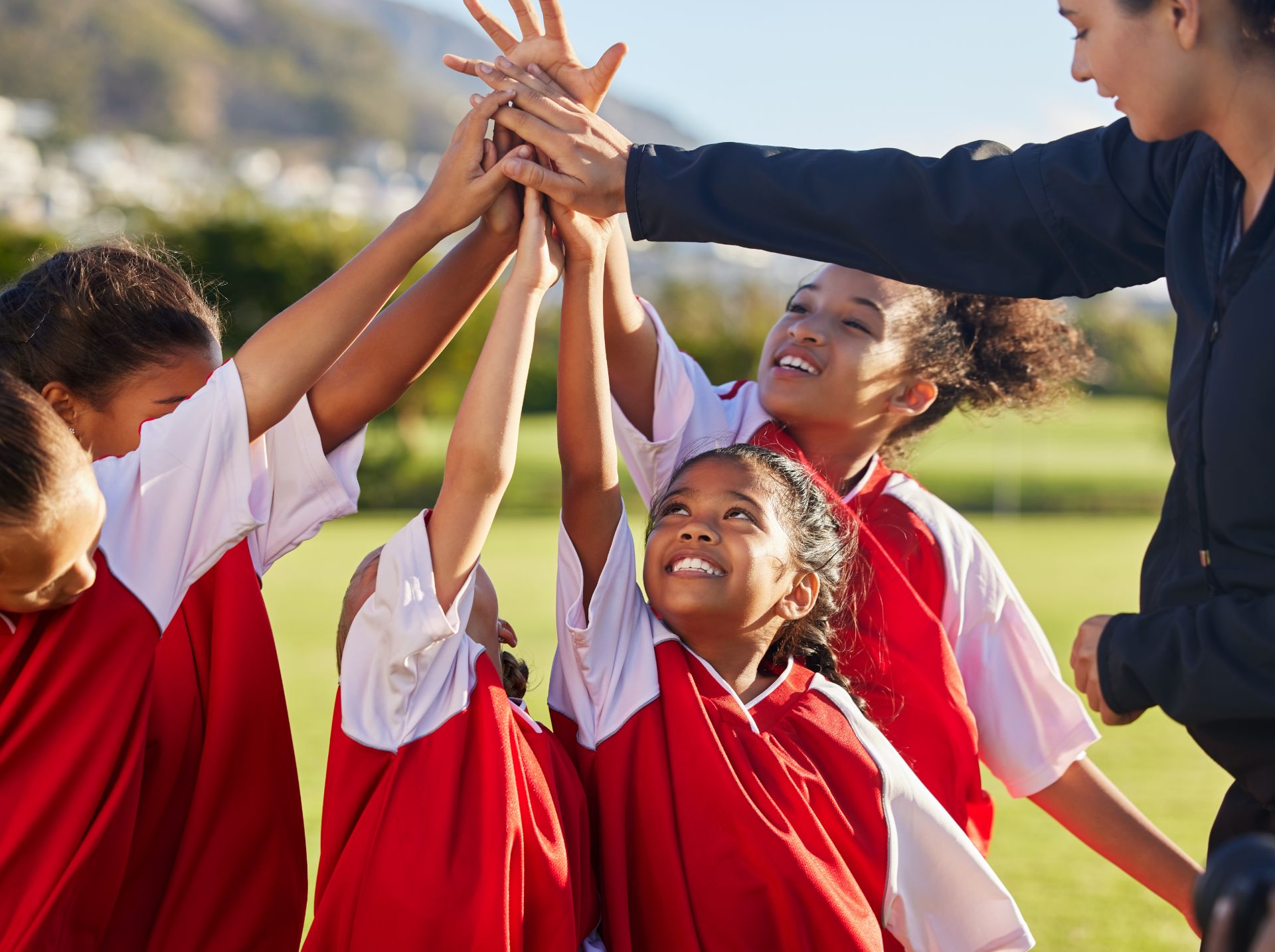 A group of young girls in red sports jerseys huddle together, reaching up with their hands in the center, outdoors on a sunny day.