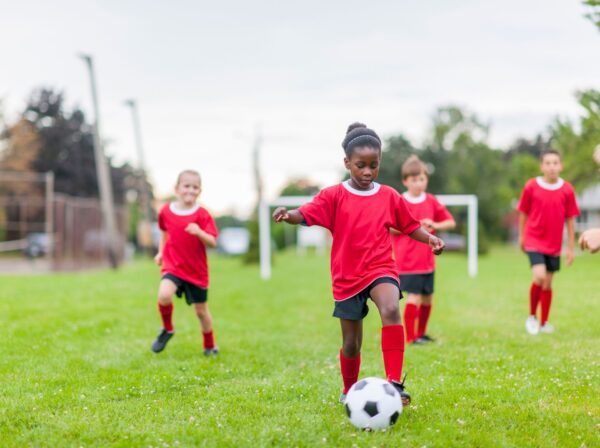 Children in red jerseys playing soccer on a green field, with one child in the foreground dribbling the ball.