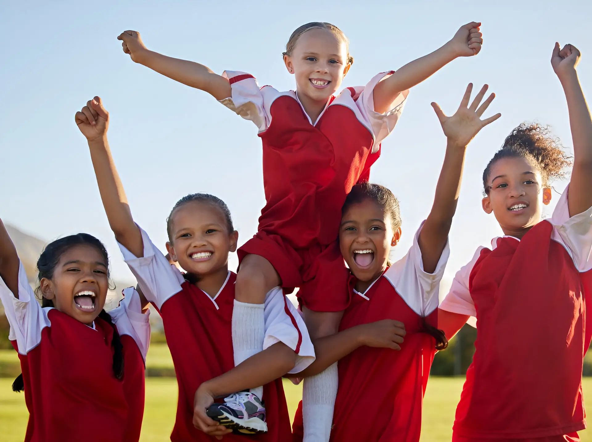 Five young girls in red sports uniforms smiling and cheering. One girl is being lifted by the others in a celebratory pose against a clear sky.
